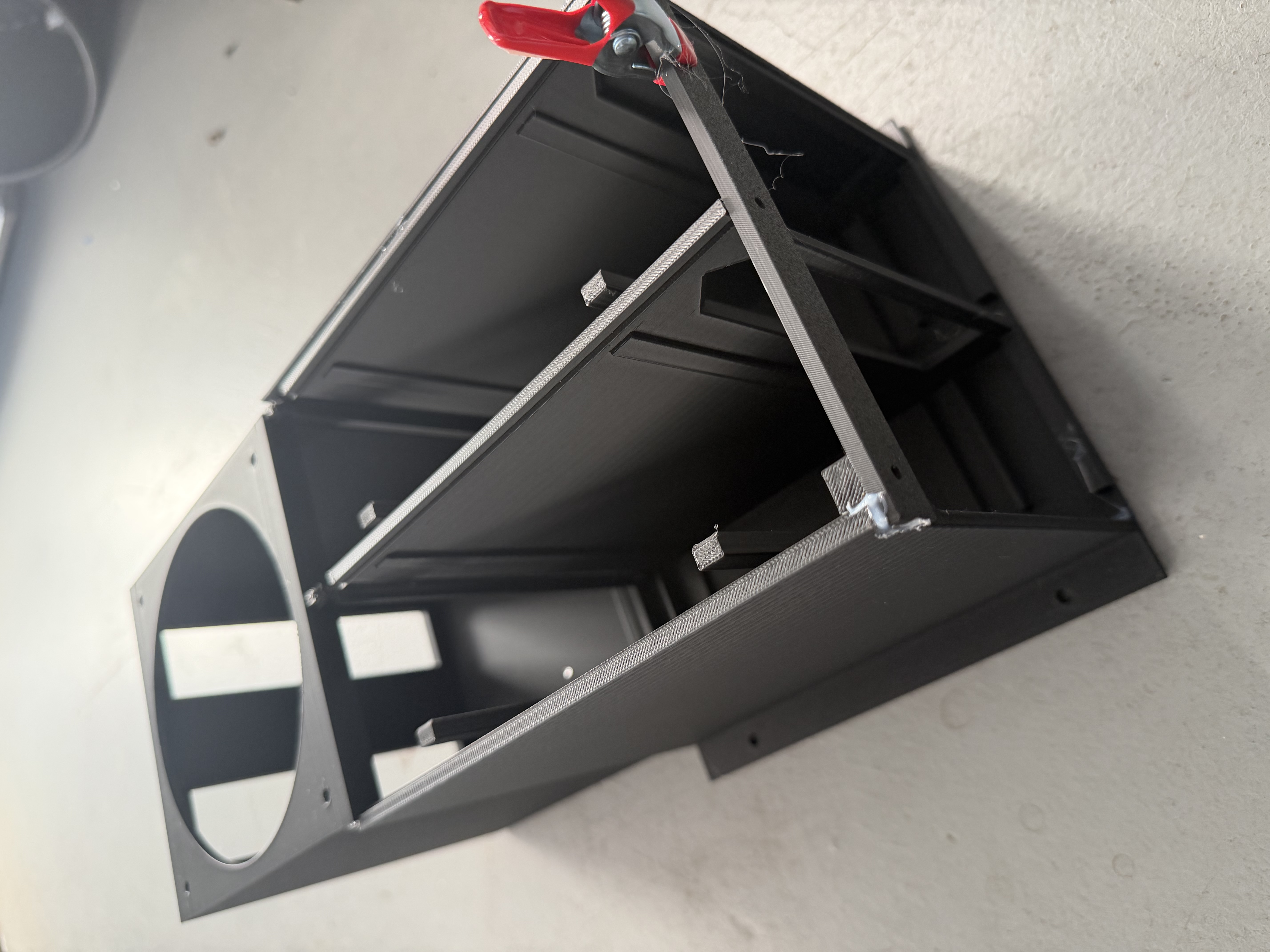 An earlier open black enclosure with internal partitions, a circular fan opening, and a red clamp on a joint.
